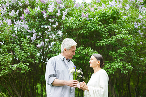 Aged man with bunch of flowers looking at his wife while both standing by blooming tree in park on summer day