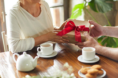 Senior female taking package with red bow on top from hands of her husband while sitting in cafe