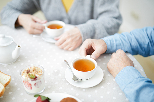 Hands of senior couple by table with cups of drinks, strawberry dessert in glass and white porcelain teapot during tea time
