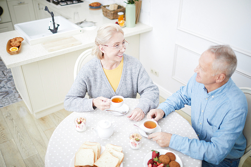 Happy grandparents sitting by served table in their kitchen, talking and having tea with desserts