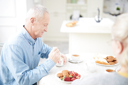 Aged man sitting by table in the kitchen and eating milk dessert with fresh strawberries