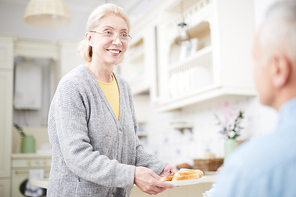 Happy grandmother bringing slices of wheat bread while serving table for tea or dinner