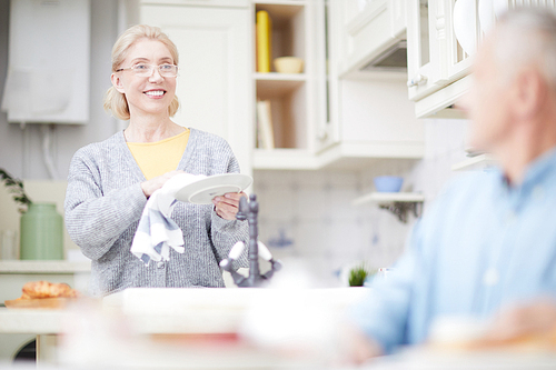 Happy aged housewife drying clean plates with towel in the kitchen and looking at her husband
