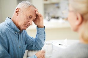 Tense or anxious senior man leaning by his hand while sitting by table with glass of water in front