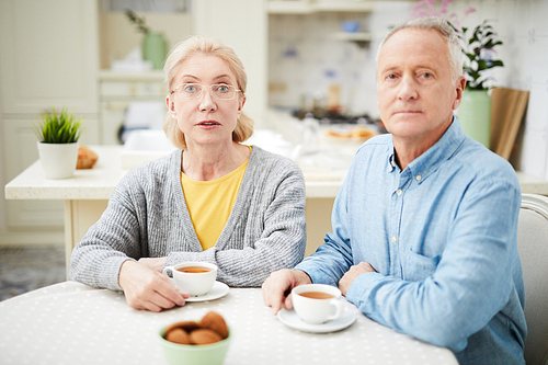 Retired couple looking at you while sitting by table and having tea with cookies in the kitchen