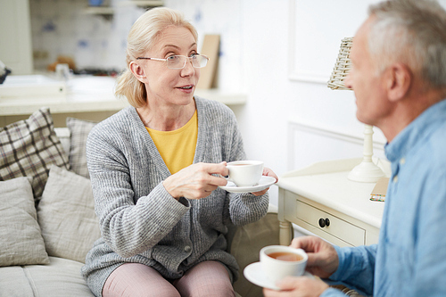 Casual senior woman with cup of tea sitting on sofa and talking to husband at home
