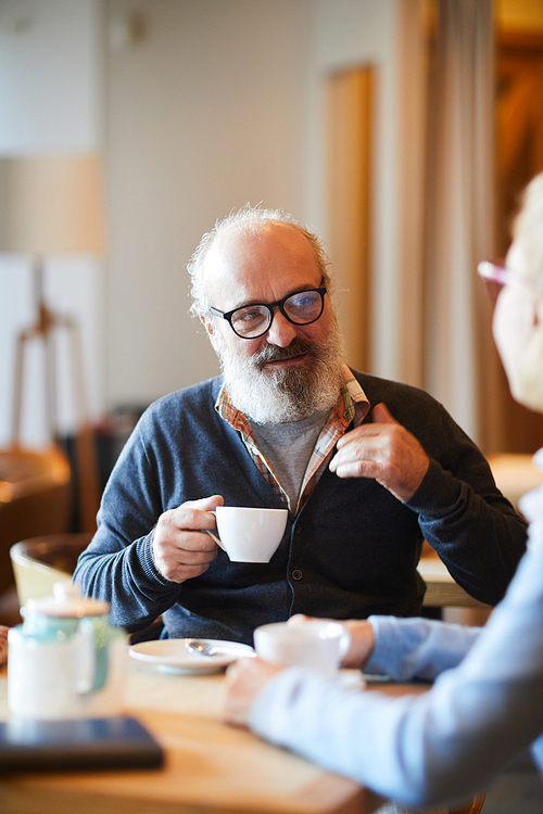 Aged confident man with grey beard having tea or coffee while sitting by table in front of his wife in cafe