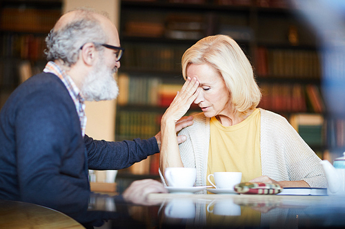 Blonde woman touching her forehead while her husband supporting her by cup of tea in cafe
