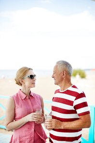 Mature spouses with drinks looking at one another while talking at summer resort on sunny day