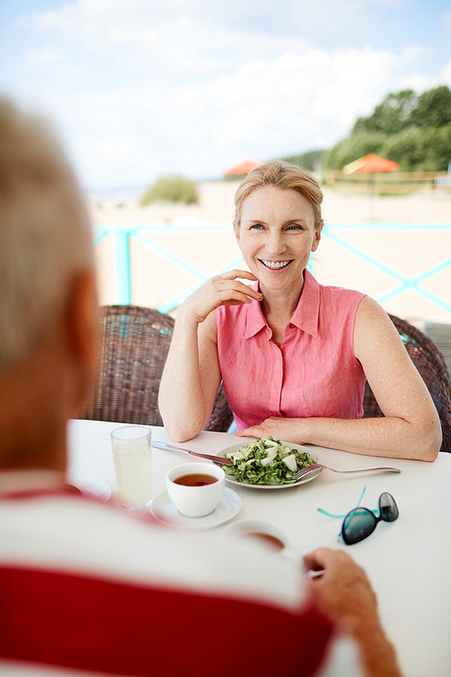 Happy mature female sitting by served table with beach behind and talking to man by breakfast