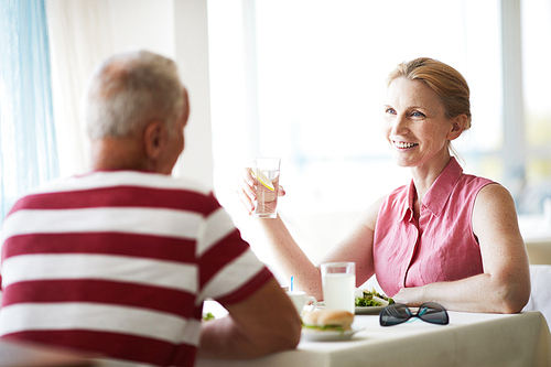 Smiling woman with glass of lemonade sitting by table in front of her husband and talking to him by lunch