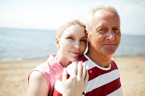 Happy and amorous mature woman standing close to her husband while both relaxing by seaside