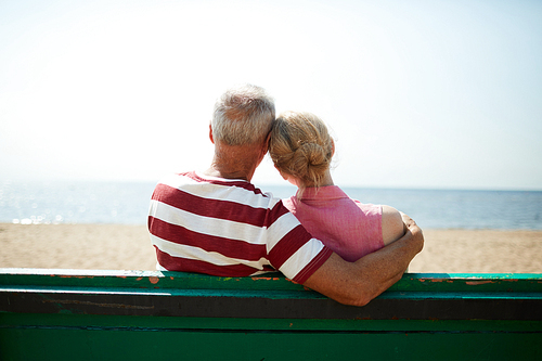 Back view of senior affectionate couple sitting on bench by waterside on hot summer day