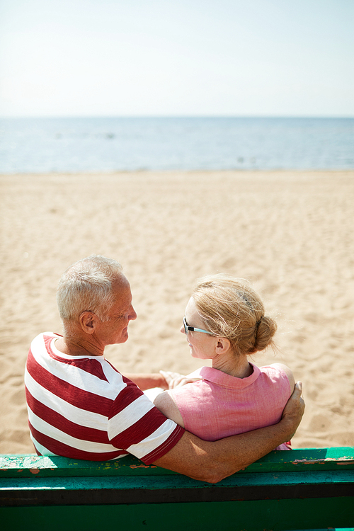 Aged couple sitting on bench, looking at each other and having talk on sandy beach by waterside