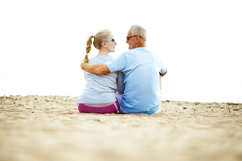 Back view of active aged spouses in sportswear sitting on sand, talking and relaxing after workout