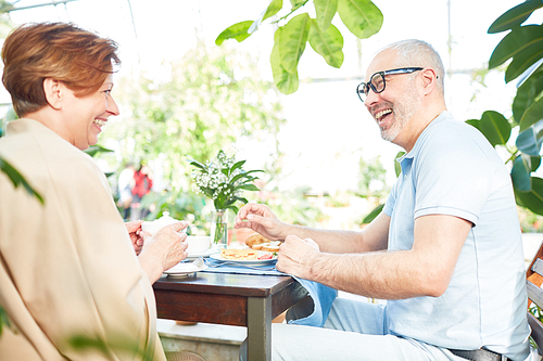 Laughing senior wife and husband sitting by served table by breakfast and having talk