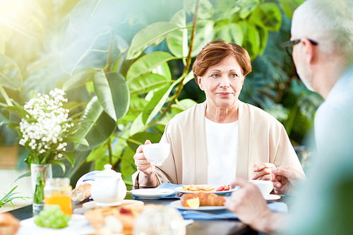 Aged wife with cup of coffee listening to what her husband telling while both sitting by served table in home garden