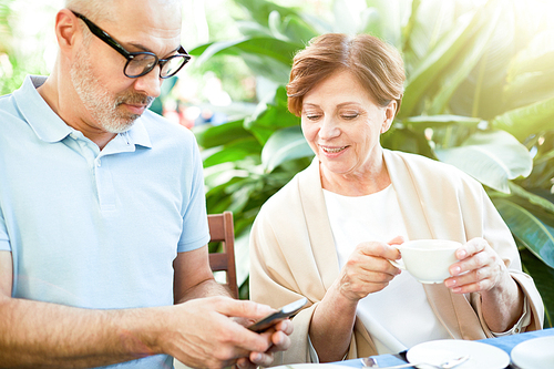 Aged man with smartphone showing message or notification to his wife by breakfast in orangery