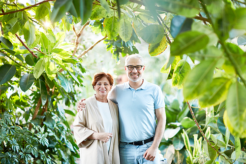 Mature affectionate couple taking walk in green garden among various trees and plants