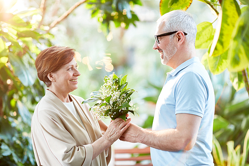 Happy senior woman taking bunch of white flowers from her husband hands on their wedding anniversay