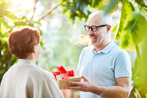 Happy senior man giving his wife box with present for their wedding anniversary during promenade in the garden