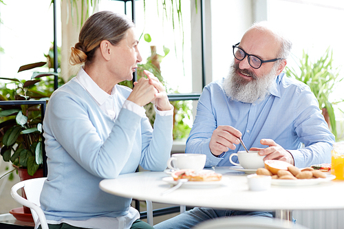 Modern senior couple sitting by table in cafe, having talk and drinking tea with cookies