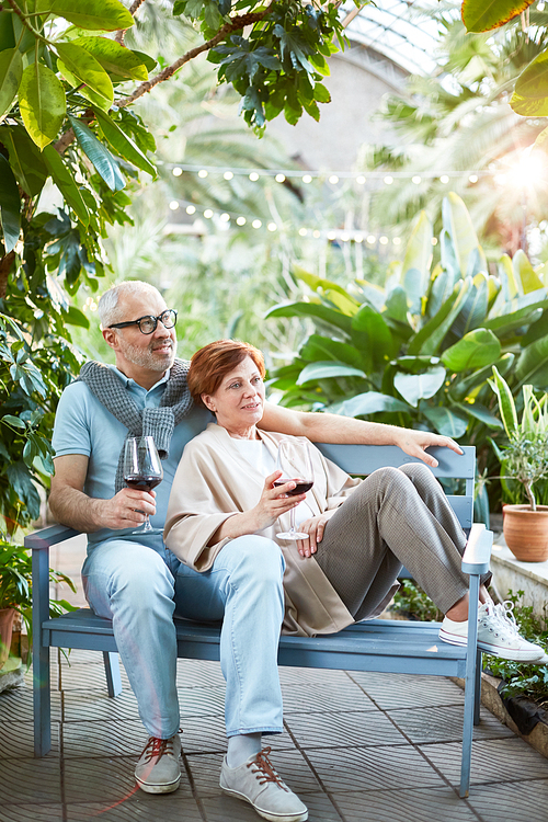 Mature couple with glasses of red wine relaxing on bench in orangery of sanatorium or tourist resort