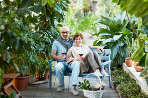 Happy and affectionate senior couple with red wine sitting on bench close to one another while having rest at tourist resort