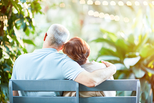 Back view of affectionate senior spouses having rest on bench in natural environment