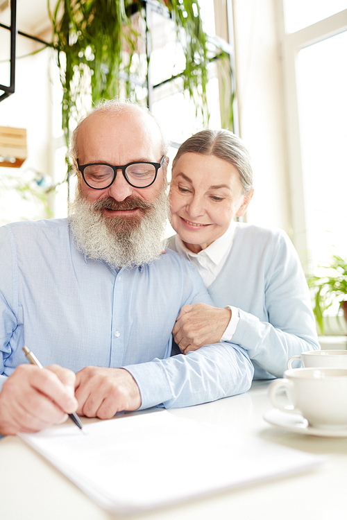 Happy mature female embracing her husband signing important paper or agreement