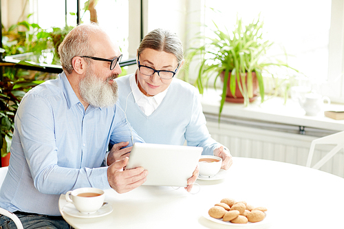 Contemporary senior couple with tablet and earphones talking through video-chat while sitting in cafe by cup of tea with cookies