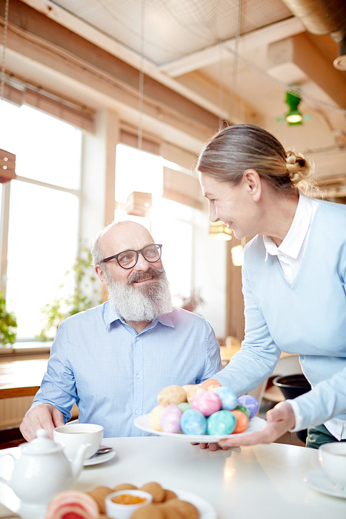 Cheerful senior man looking at his wife bringing plate with heap of Easter eggs