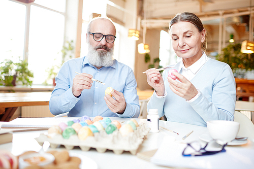 Senior couple sitting by table and painting eggs for Easter festivity with special paints