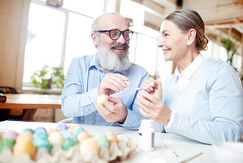 Cheerful mature couple discussing colors of paints for eggs while preparing for Easter