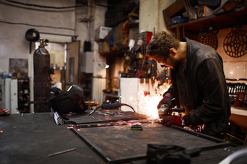 Serious concentrated young worker in leather jacket sanding metal part while producing metal frame in smithery