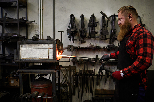 Serious brutal blacksmith with long beard heating steel bar in small furnace and holding hammer while standing in workshop