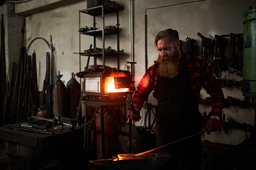 Bearded professional blacksmith with hammer forging hot end of iron workpiece on anvil
