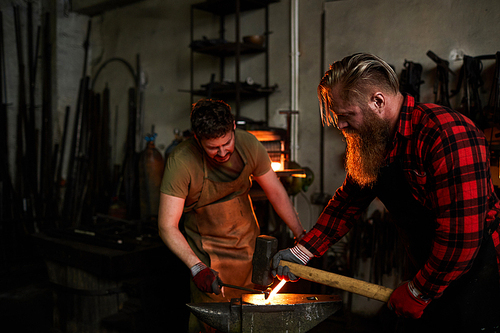 Team of brutal blacksmiths in workwear shaping metal together: bearded man hammering heated metal held by his coworker in factory shop
