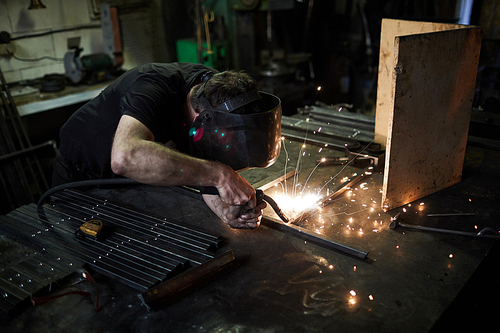 Concentrated craftsman in mask using welding torch while producing metal frame and joining wrought iron parts in workshop