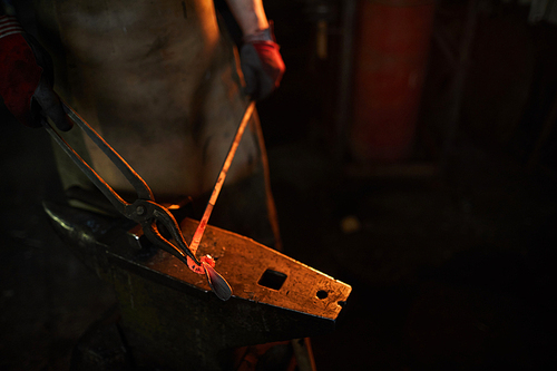 Close-up of unrecognizable blacksmith in apron shaping heated metal plank with tongs while working at anvil in dark workshop