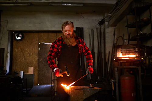 Bearded blacksmith in flannel and apron standing by anvil and forging hot molten metal