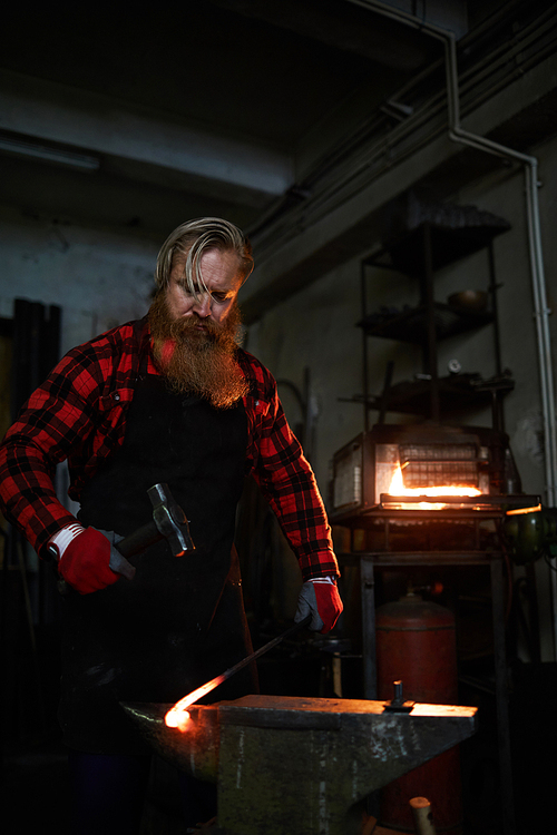Serious brutal handsome blacksmith in checkered shirt and apron forging wrought iron bar on anvil and beating it with hammer in dark workshop
