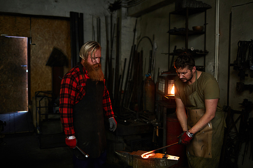 Skilled long-bearded blacksmith in apron teaching young worker to forge metal and controlling his work in dark workshop