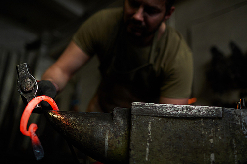 Close-up of busy manual worker forging heated metal with hammer on anvil while working in dark workshop