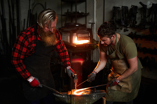 Two professionals in workwear leaning over anvil and forging molten metal workpiece
