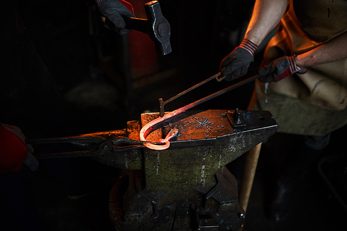 Close-up of unrecognizable blacksmiths producing metal part together: one of them holding heated bar and fire iron while his colleague hammering detail with hand tools