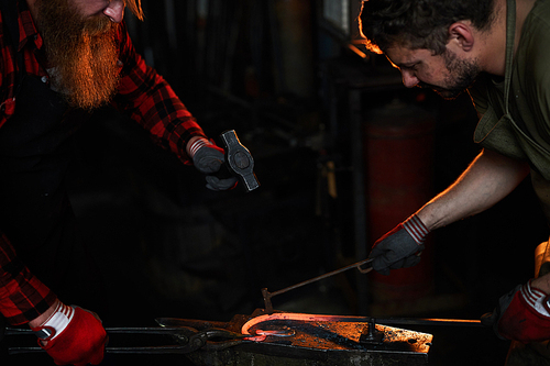 Concentrated team of blacksmiths shaping heated metal on anvil: worker using fire iron and fixing heated metal bar while other man beating it with hammer