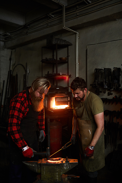 Serious confident blacksmith in apron showing concentrated coworker how to shape heated metal with hammer