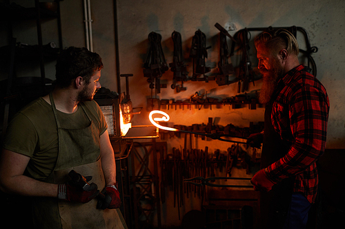 Blacksmiths at work: serious long-bearded smith getting heated metal bar out of furnace and checking it while working with assistant in workshop