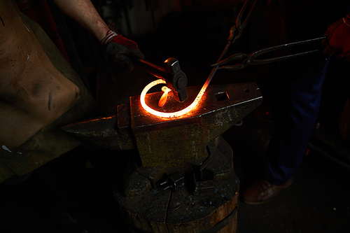 Close-up of unrecognizable blacksmiths hammering heated iron bar on anvil while making twisted shape for fence